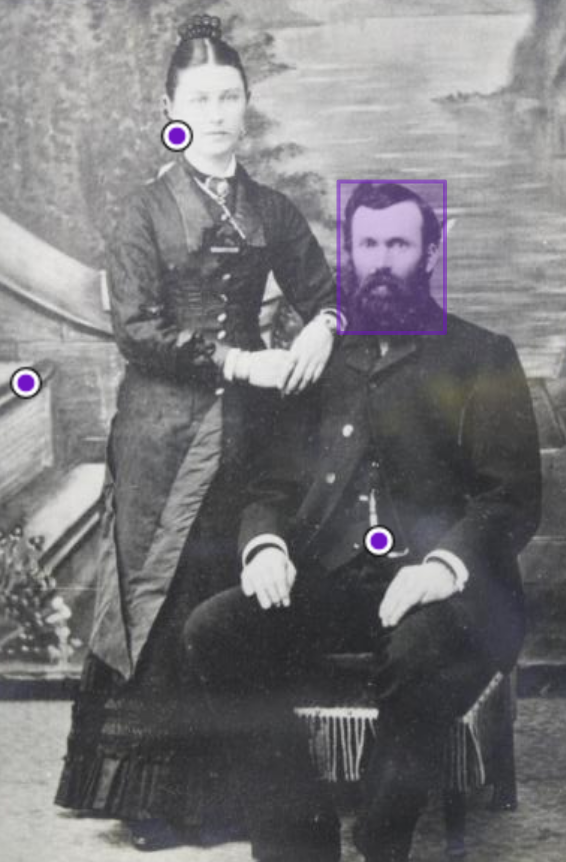 Black and white photograph of man seated with woman standing next to him. The photo has purple dots indicating it has been annotated.